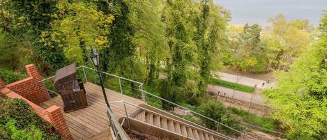 A wooden staircase leading to a deck with outdoor seating, surrounded by lush greenery and overlooking a body of water.