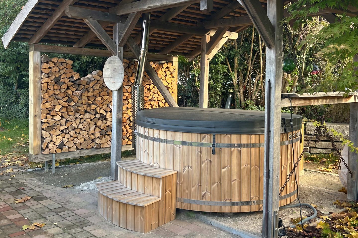 Een houten jacuzzi met trap, een overdekt gedeelte en een stapel haardhout.