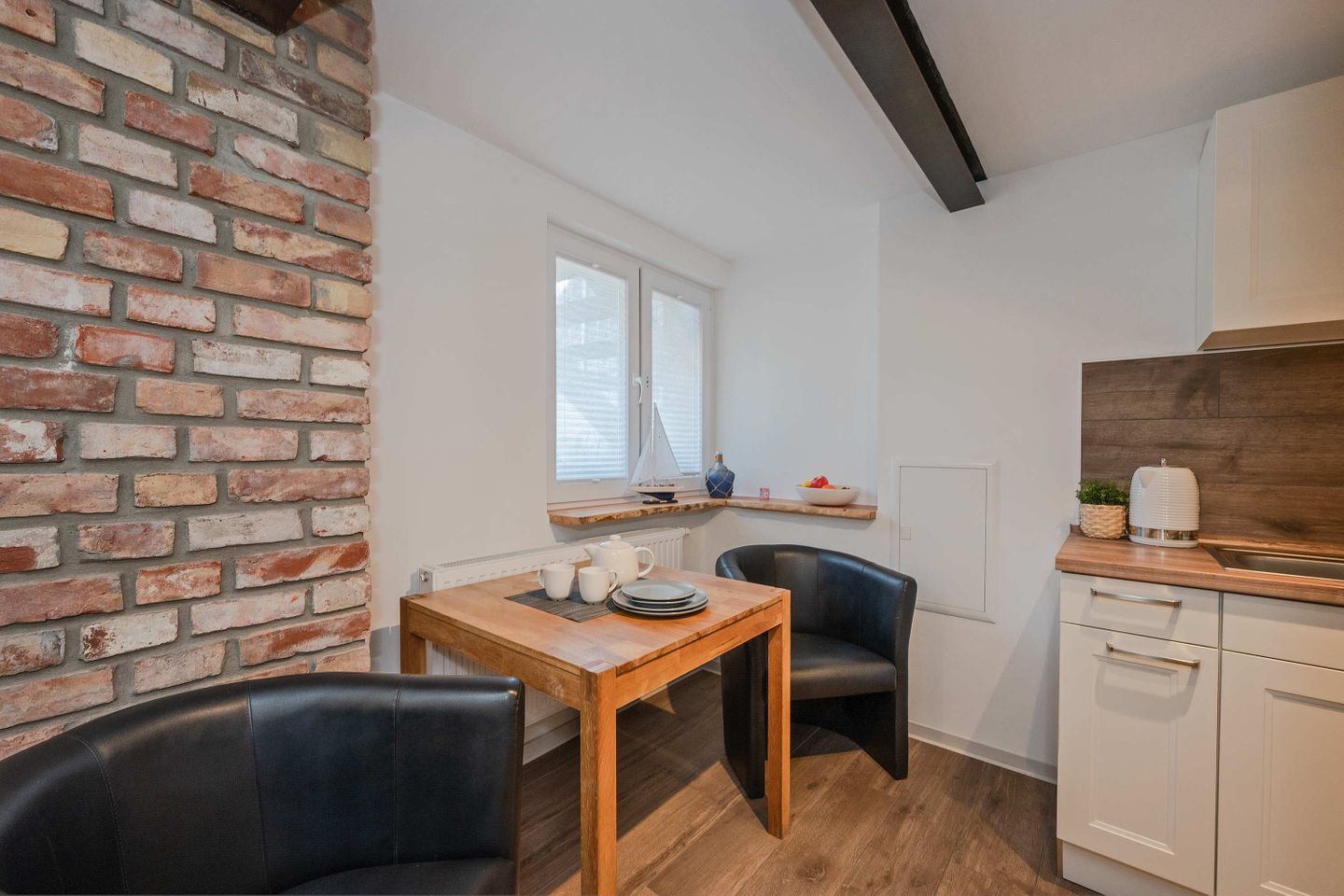 A compact kitchen with a brick wall, a wooden table, two chairs, a window, and a white cabinet.