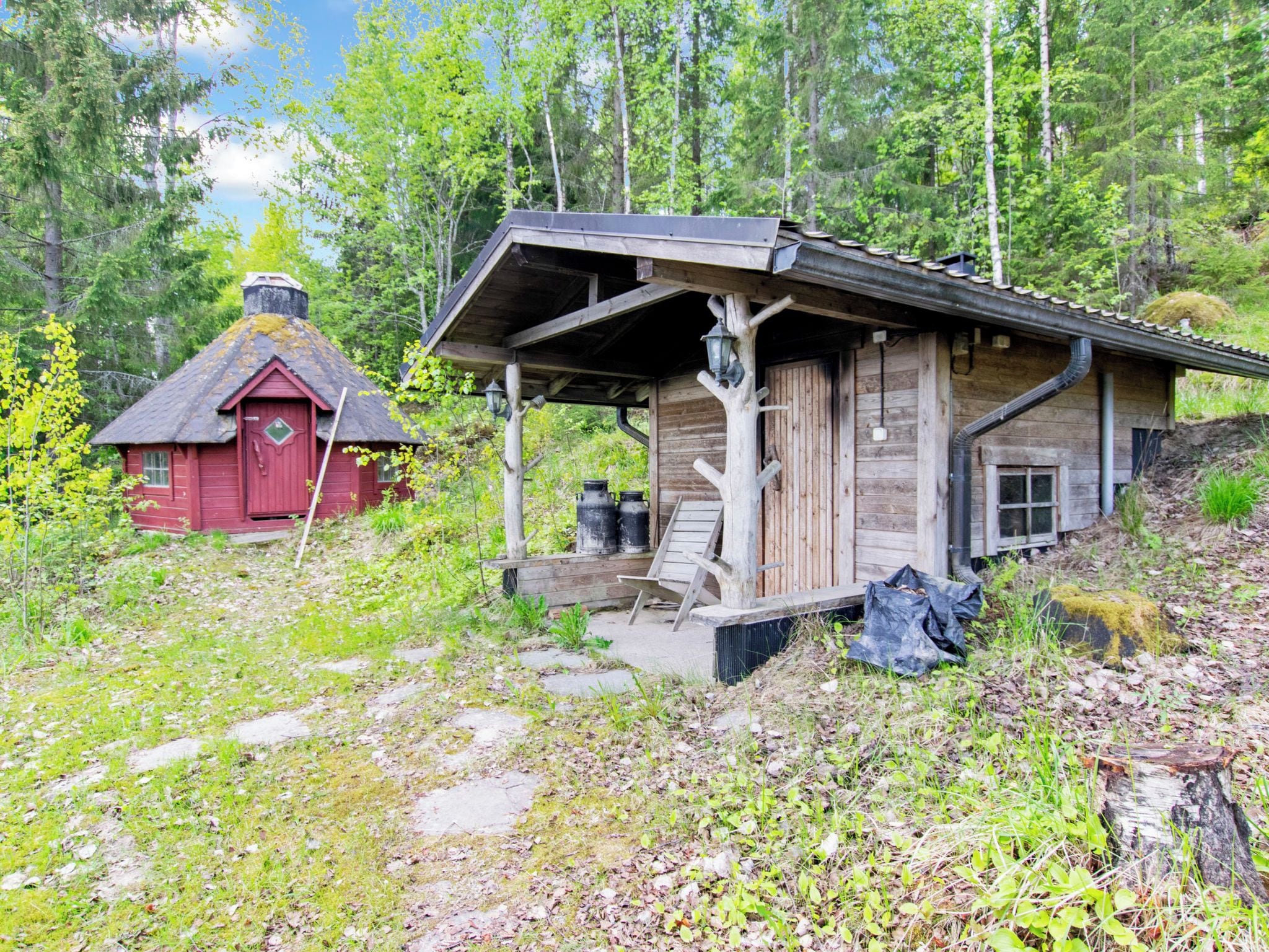 A rustic cabin with a porch, a red shed, and a forested backdrop.
