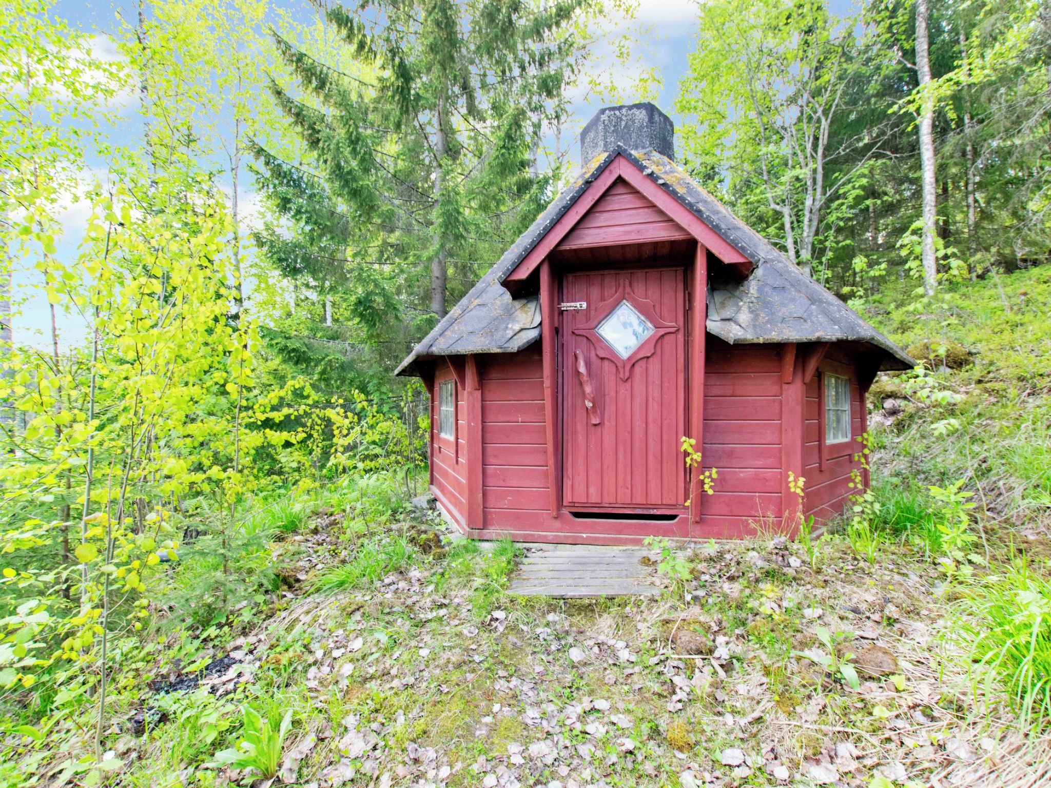 Pieni, punainen puumökki, jonka katto on erittäin jyrkkä, ja jonka ympärillä on tiheä metsä.