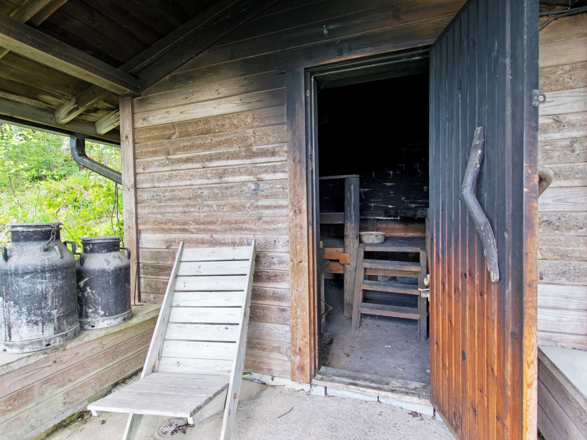 Une cabane en bois avec une rampe en bois menant à une porte, un baril en métal et un banc en bois.