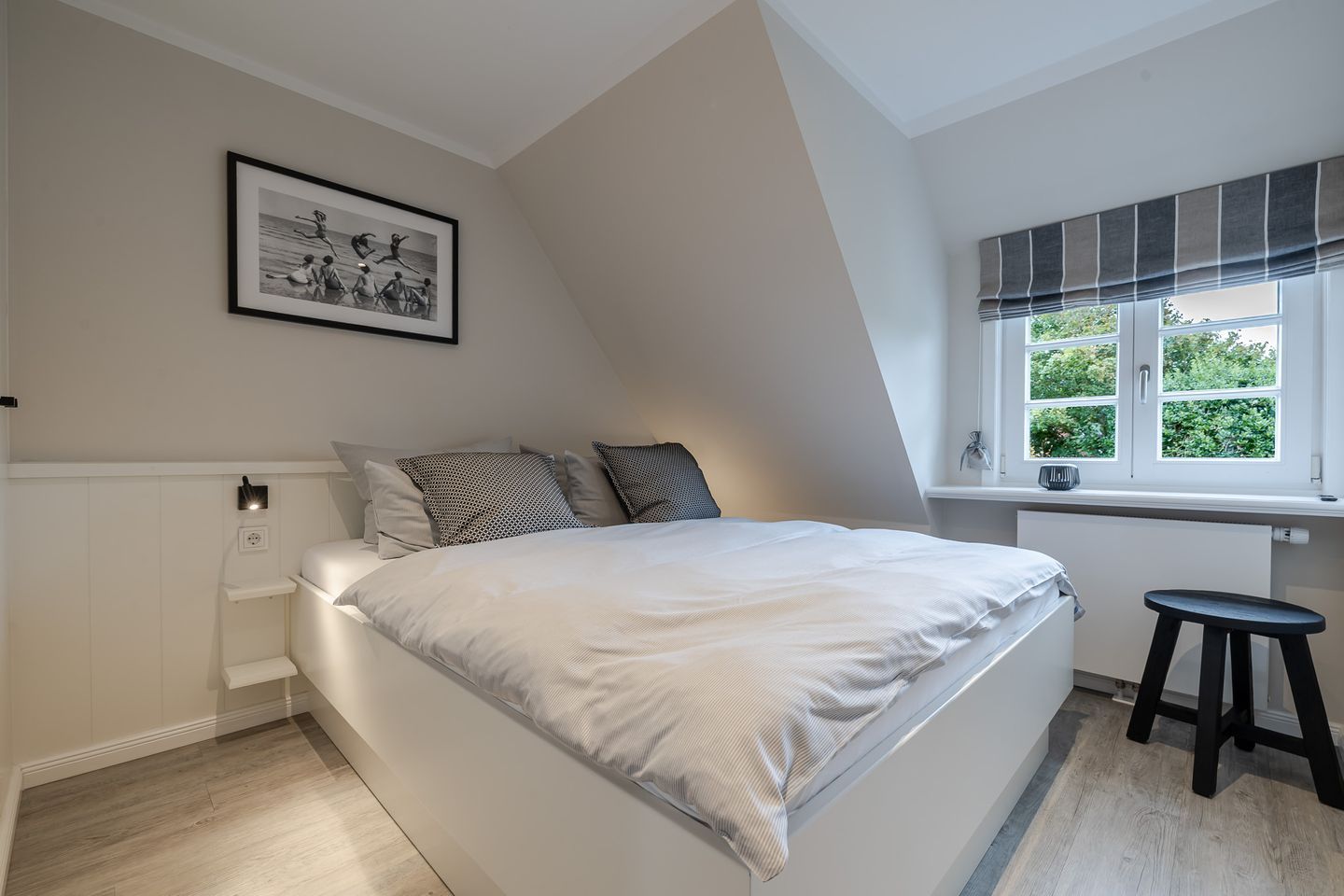A bedroom with a bed, a window with curtains, a picture on the wall, and a stool.