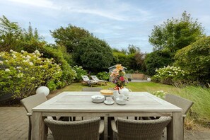 A garden patio with a table set for tea, surrounded by lush greenery and a wooden fence. - Hedigen Hüs - Our Hedigen Hüs impresses with its consistent furnishings in the Sylt style. (Sylt)