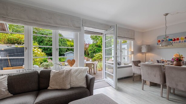 A modern living room with a grey sofa, a dining area with a table and chairs, and large windows with a view of greenery outside. - Hedigen Hüs - Our Hedigen Hüs impresses with its consistent furnishings in the Sylt style. (Sylt)