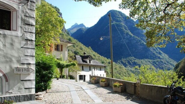 Una calle estrecha con un cartel que dice "Valle di Prato", un edificio de fachada de piedra y una montaña al fondo.