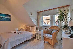 Un dormitorio con cama, una silla, una mesita de noche, una ventana con cortinas y una planta.