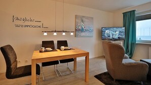 A modern dining area with a wooden table, four chairs, and pendant lighting. A television is mounted on the wall to the right, and a painting is displayed above it.