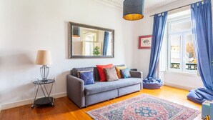 A living room with a grey sofa, a patterned rug, a lamp, and a large window with blue curtains. - Cozy Lisbon Stay - 2 Min Walk from Av. Liberdade by Lisbeyond (Lisboa)