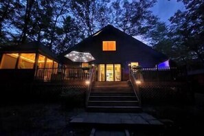 Une cabane avec une véranda, un escalier menant à l’entrée et une fenêtre dont les lumières sont allumées.