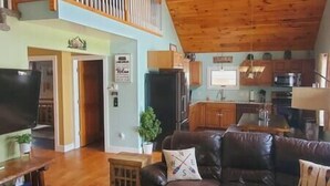 A spacious living area with a wooden ceiling, a dark leather sofa, a television, and a kitchen in the background.