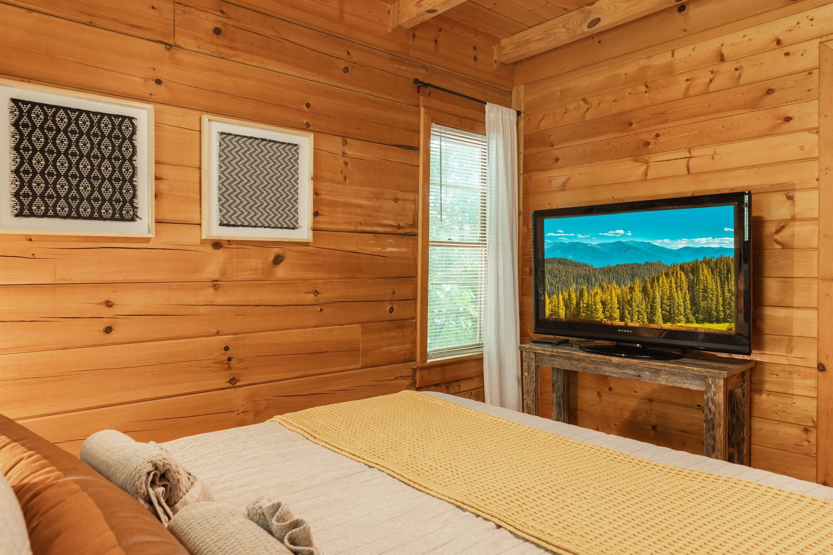 A wooden cabin room with a bed, a television on a wooden stand, and two framed pictures on the wall.