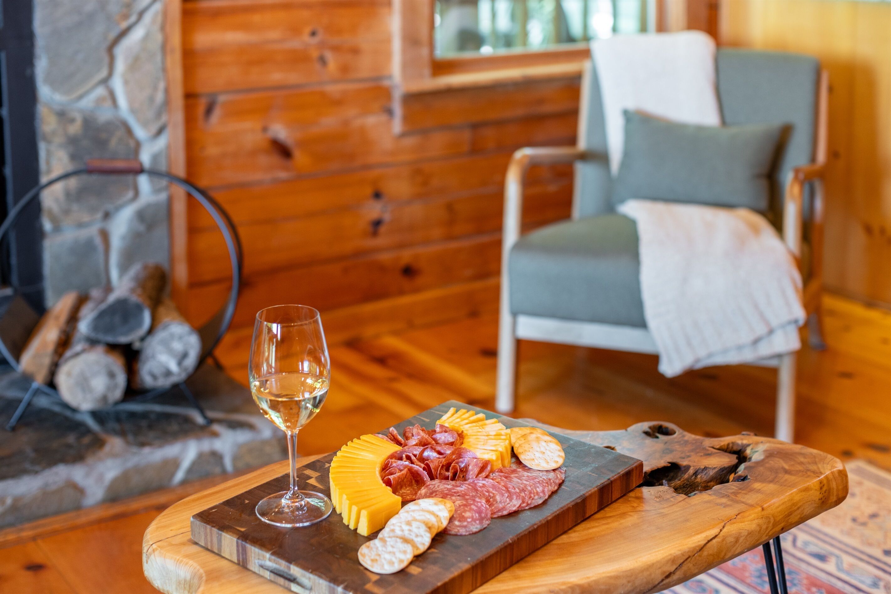 A wooden dining table with a charcuterie board, a glass of white wine, and a stone fireplace in the background.
