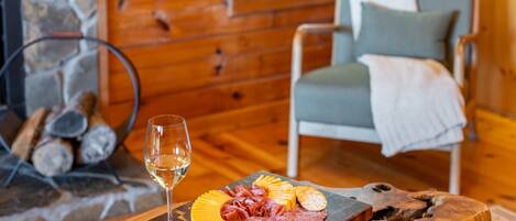 Una mesa de comedor de madera con una tabla de embutidos, una copa de vino blanco y una chimenea de piedra al fondo.