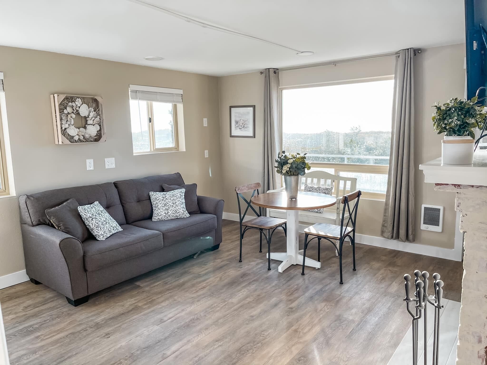 Ein modernes Wohnzimmer mit einer grauen Couch, einem runden Holztisch und einem Balkon mit Aussicht.