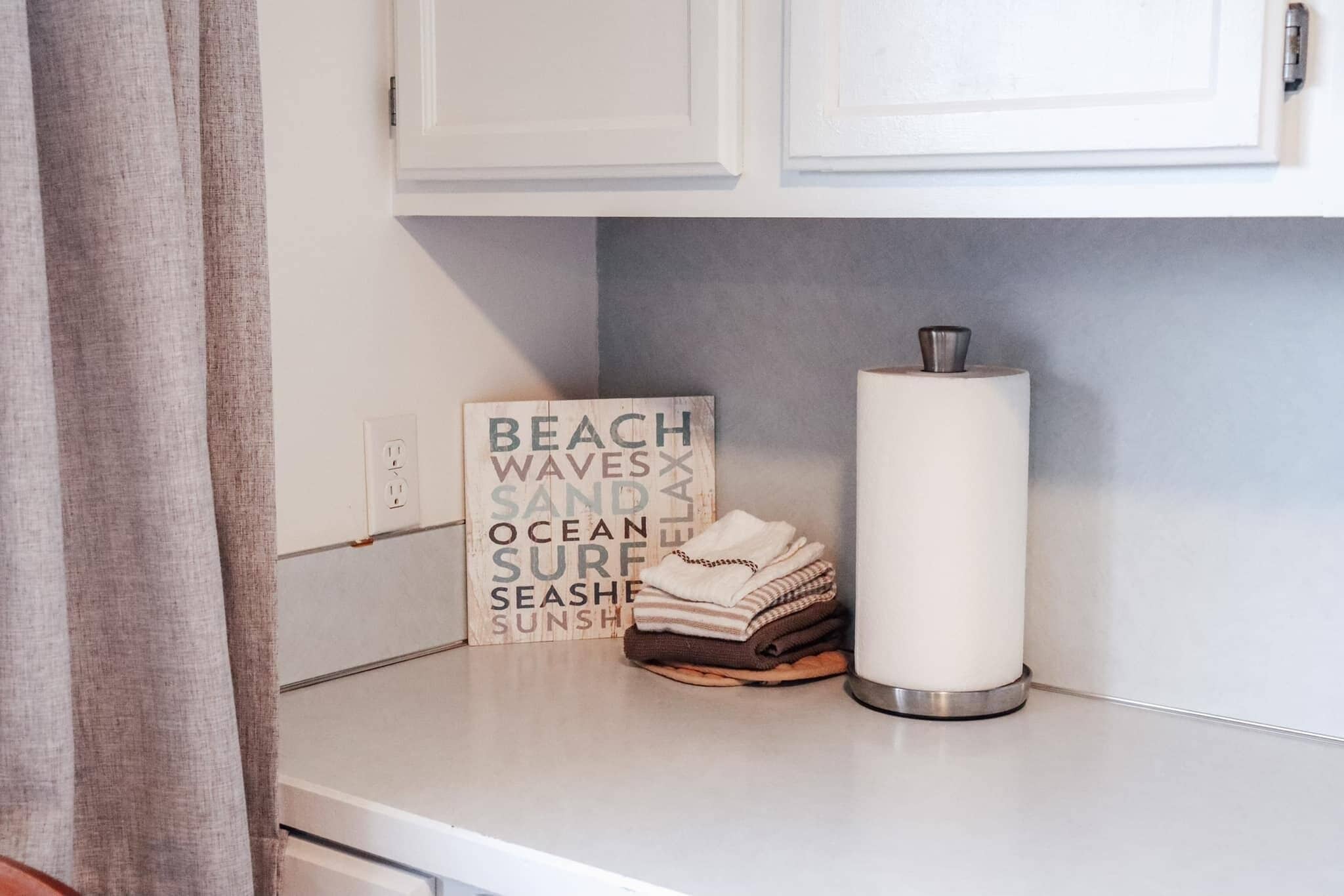 A bathroom with a white countertop, a towel rack with folded towels, a decorative sign with beach-related words, and a cylindrical container.