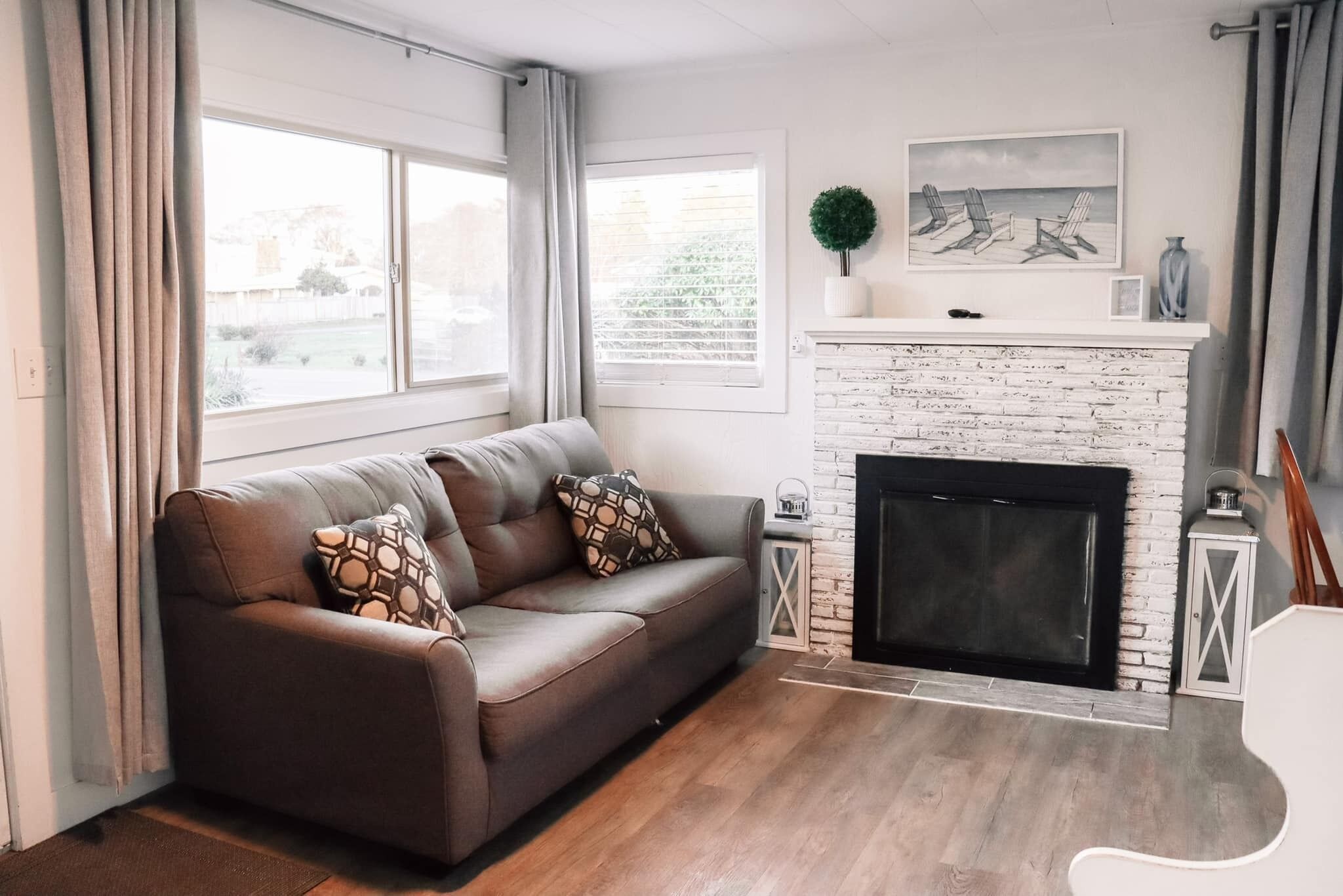 A living room with a brown leather sofa, a white brick fireplace, and a large window with curtains.