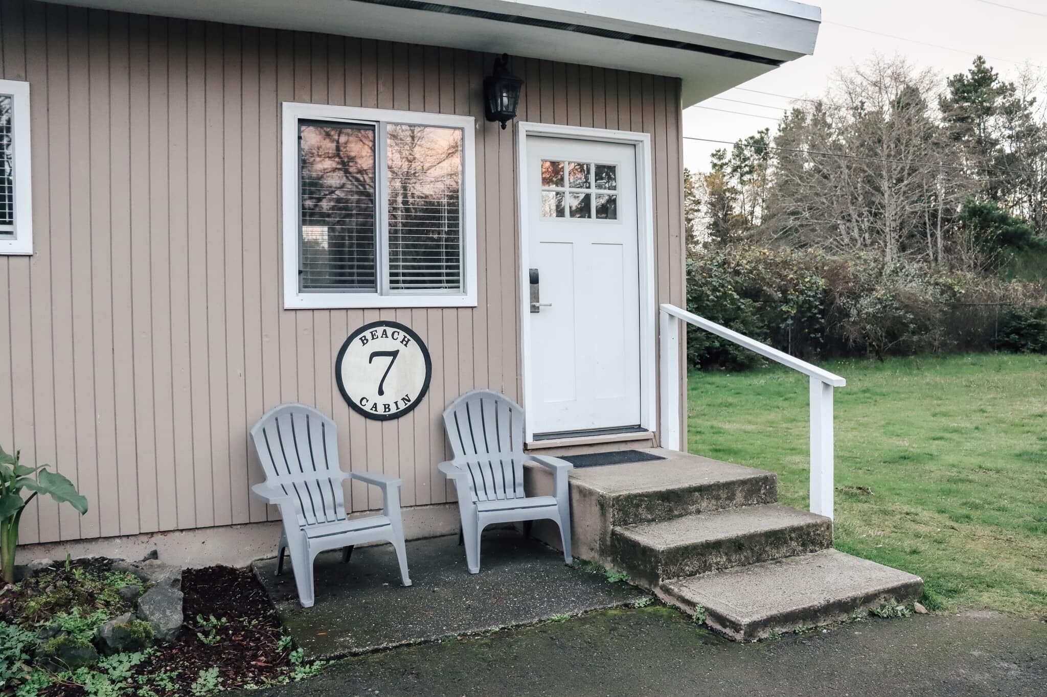 A cabin with two Adirondack chairs outside, a white door, and a number '7' sign on the wall.