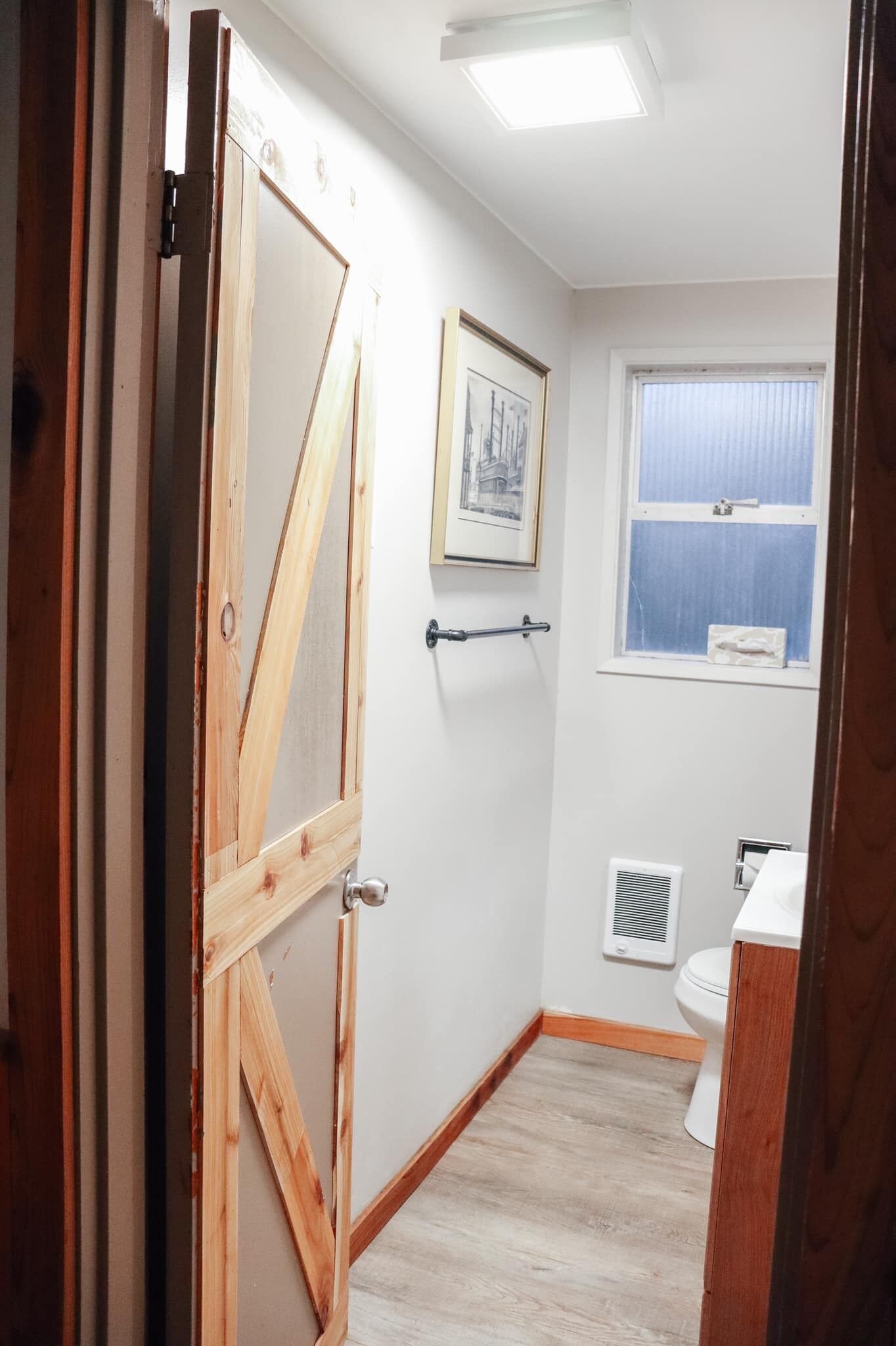 A bathroom with a wooden door, a framed picture on the wall, a window with blinds, a toilet, and a vent.