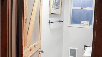 A bathroom with a wooden door, a framed picture on the wall, a window with blinds, a toilet, and a vent.