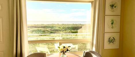 A room with a large window offering a view of a coastal landscape, a round table with chairs, and three framed pictures on the wall.