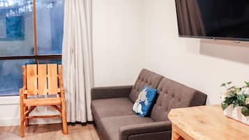 Uma sala de estar moderna com um sofá cinzento, uma cadeira de madeira, uma mesa de madeira e uma televisão grande fixada na parede.