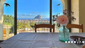 Uma mesa com um vaso de flores, uma cadeira e vista para uma cidade costeira com pássaros voando.
