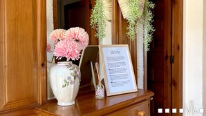 Balcão de recepção de madeira com um vaso de flores cor-de-rosa, uma placa de boas-vindas emoldurada e plantas penduradas.