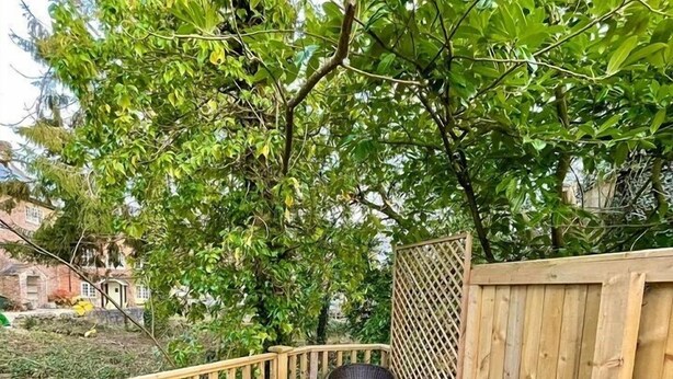 Una terraza de madera con mobiliario de mimbre y una mesa de vidrio, rodeada de abundante follaje y una valla de madera.