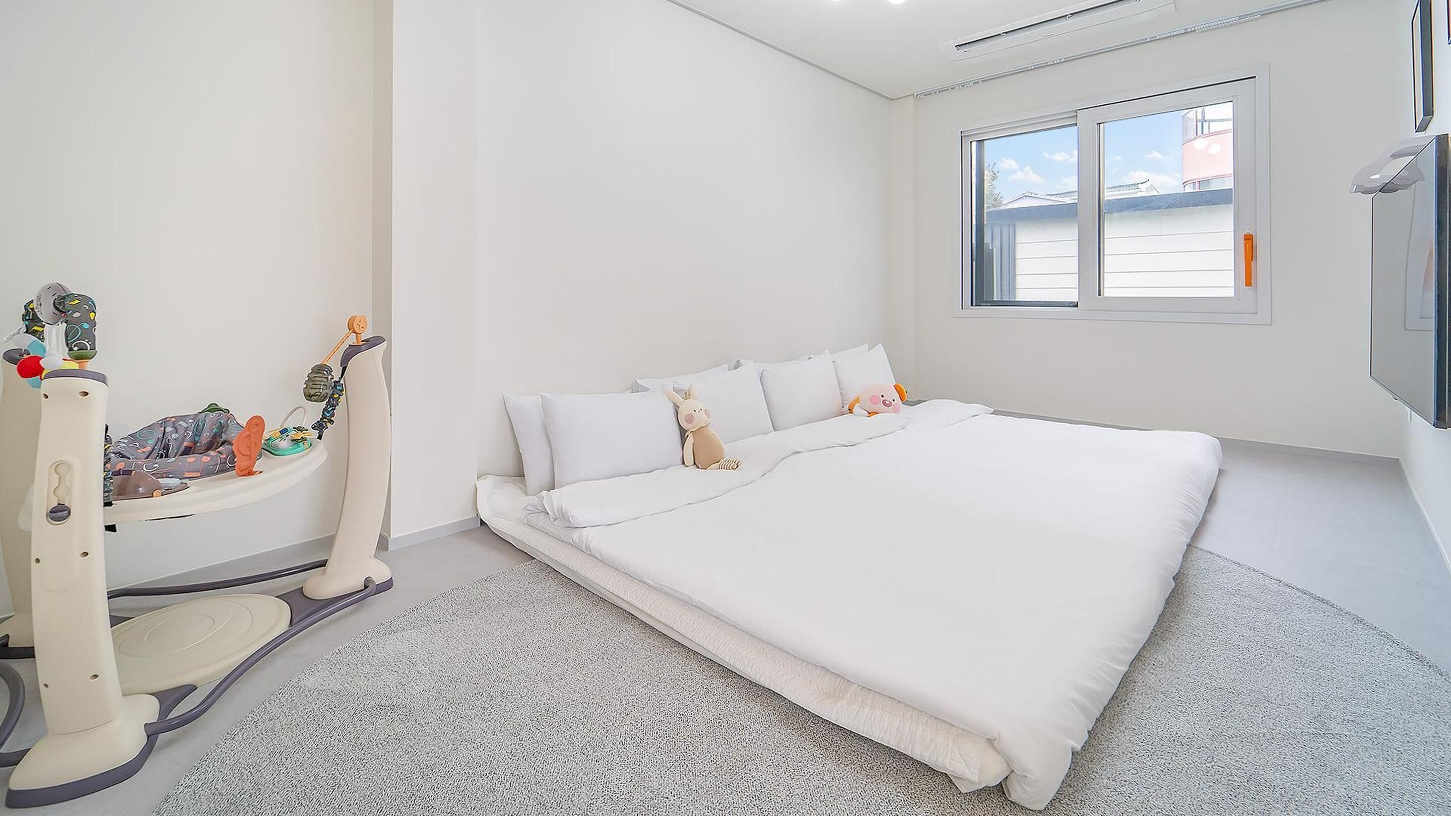 A bedroom with a large bed, a white wall, a window, and a crib with toys.