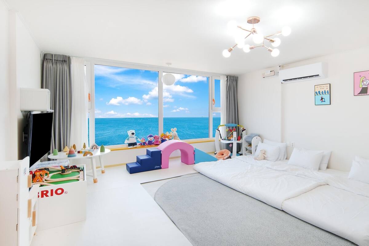 Ein Schlafzimmer mit großem Fenster zum Meer, einem Bett, einem Schreibtisch mit Spielzeug und einem Babybett.