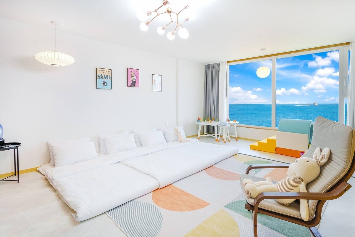 Ein geräumiges Zimmer mit einem großen Bett, einem Sessel mit Kuscheltier, einem Schreibtisch mit Spielzeug und Meerblick.