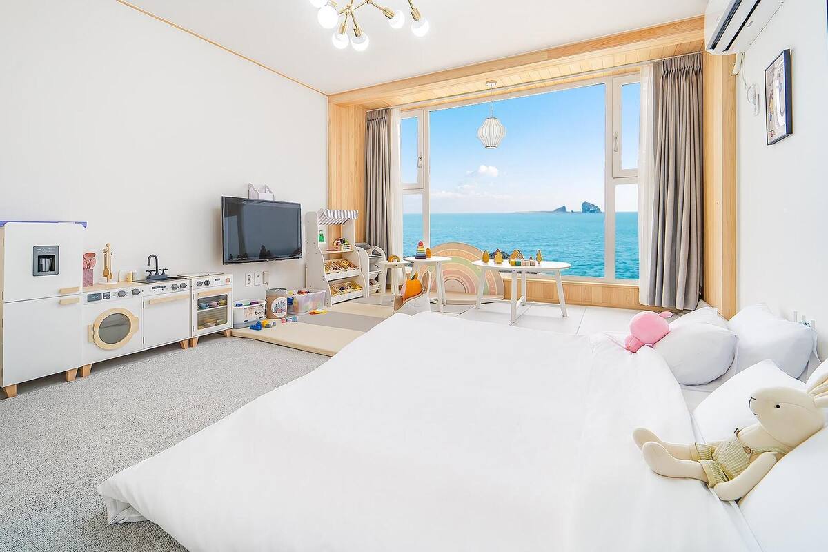 Ein Hotelzimmer mit einem großen Bett, einem Fernseher, einer Küchenzeile, Meerblick und einem Vorhangfenster.