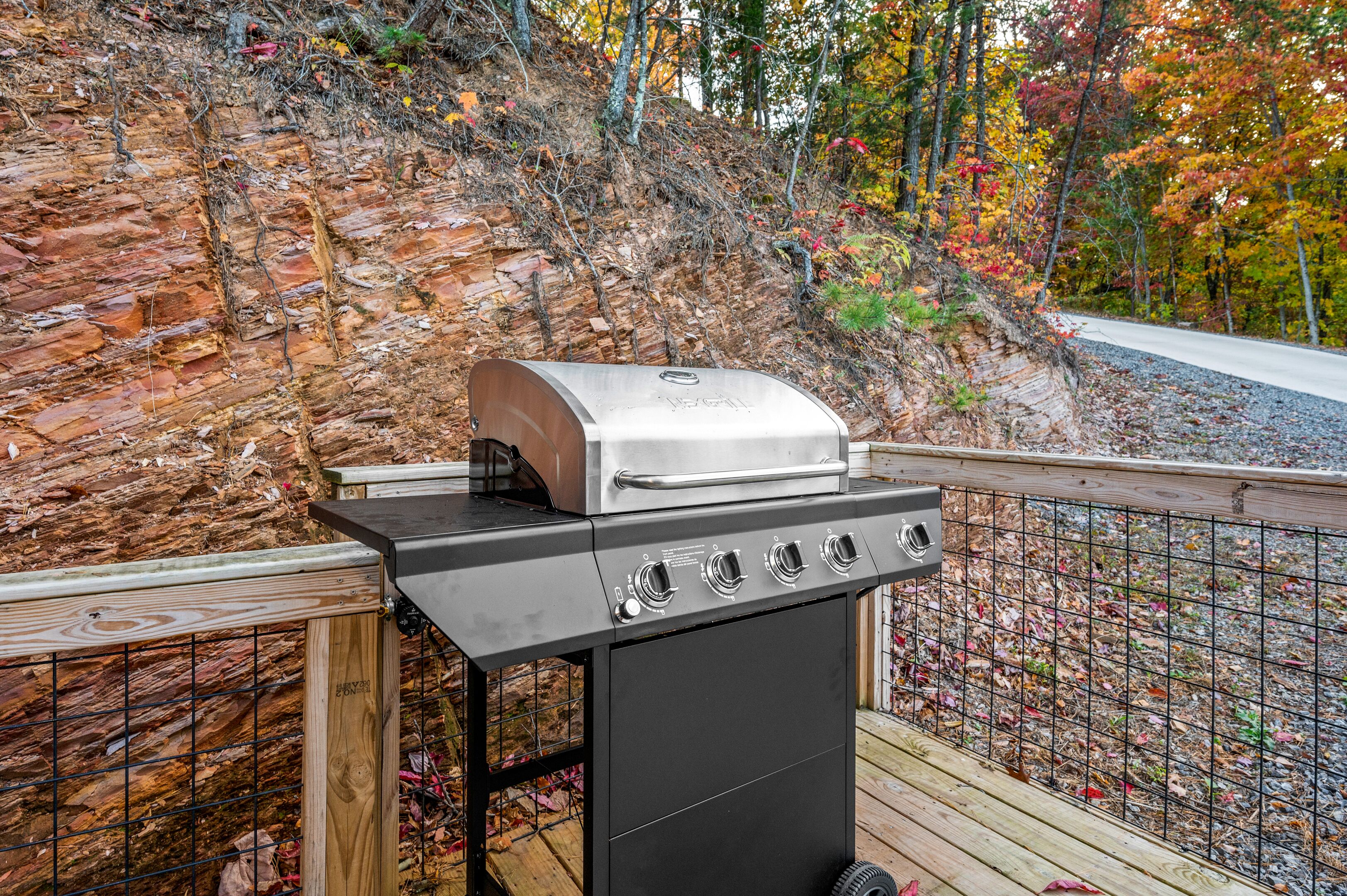 A barbecue grill on a wooden deck with a stone wall and autumn foliage in the background.