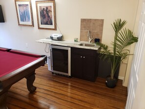 A pool table, a built-in mini fridge, a sink, and a potted plant in a room with wooden flooring and tiled walls.