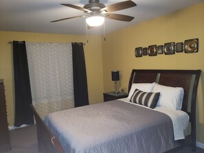 A bedroom with a bed, a ceiling fan, a nightstand with a lamp, and a wall-mounted decoration.