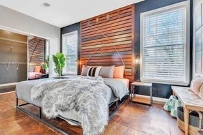 A modern bedroom with a wooden headboard, a large bed with a fluffy blanket, a bedside table, and a window with blinds. - Eva's Compound | 3-Unit Estate | Near Mercedes-Benz Stadium (Atlanta)
