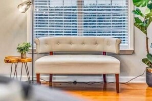 A room with a tufted bench, a small wooden side table with a plant, and a window with blinds.