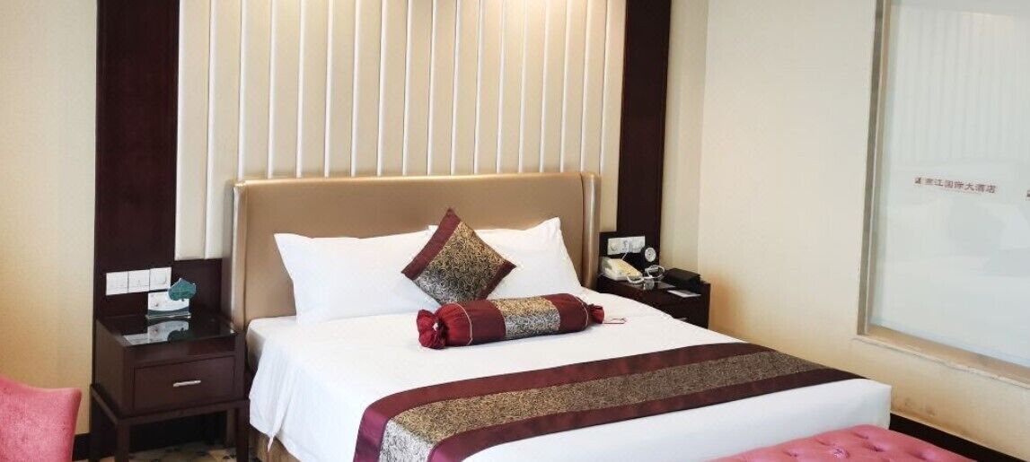 A hotel room with a bed, a nightstand, a bedside table, a lamp, and a window with blinds.