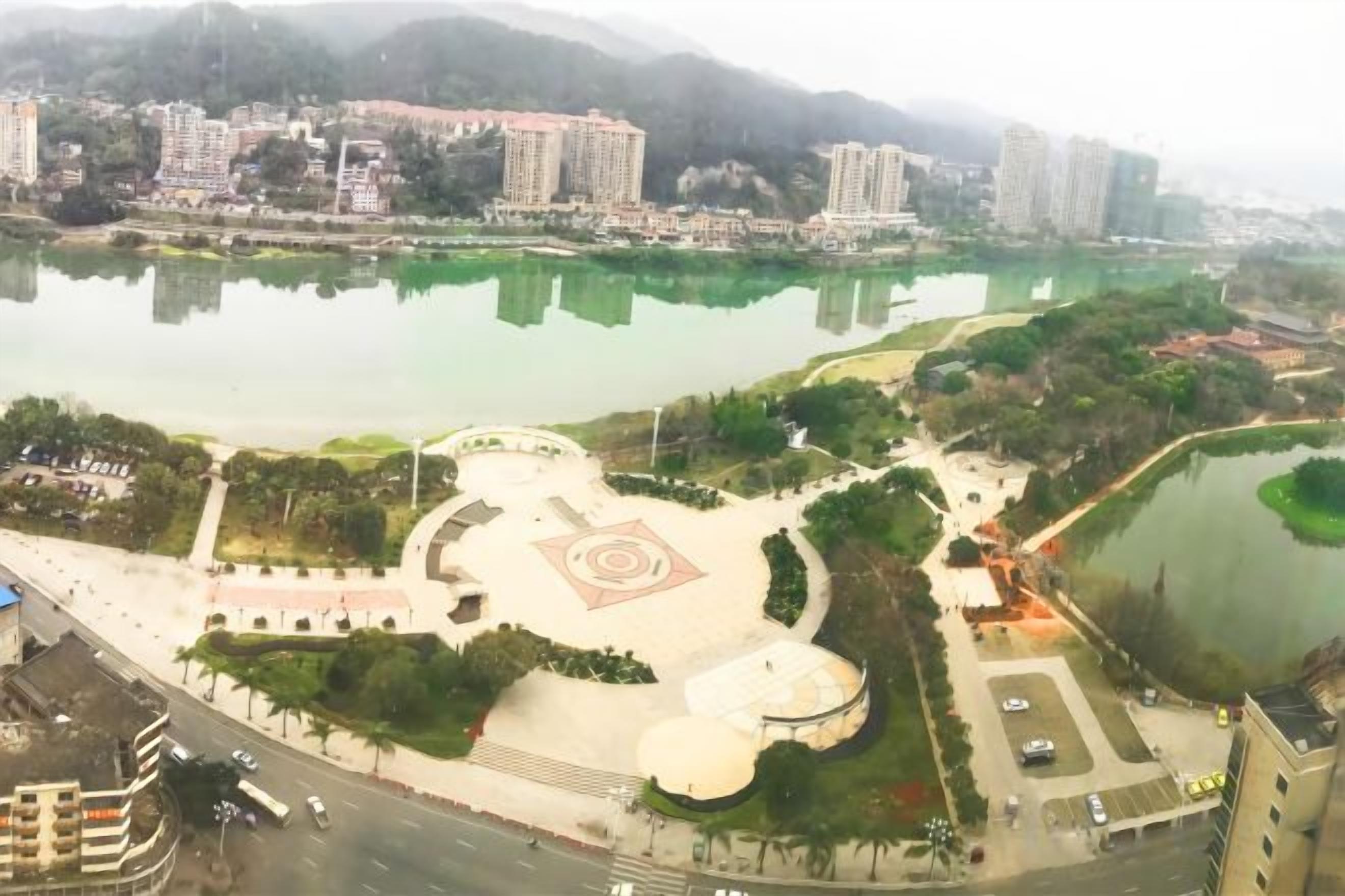 Un parque con una plaza circular central, un lago y edificios circundantes.
