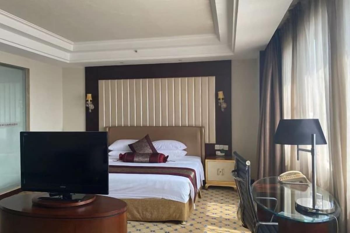A hotel room with a bed, a television, a bedside table, a lamp, and a window with curtains.