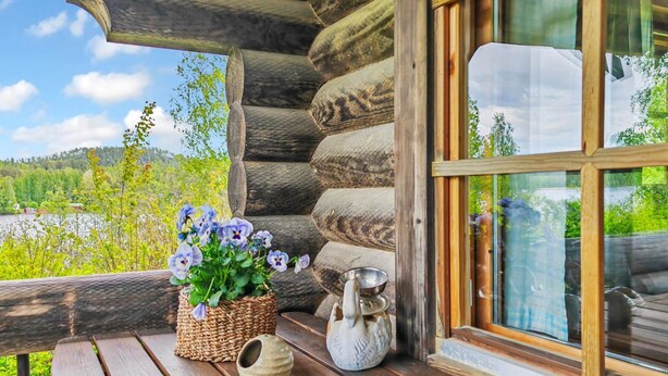 Una cabaña de madera con pared de troncos, un banco de madera, una cesta tejida con flores y una maceta de cerámica.