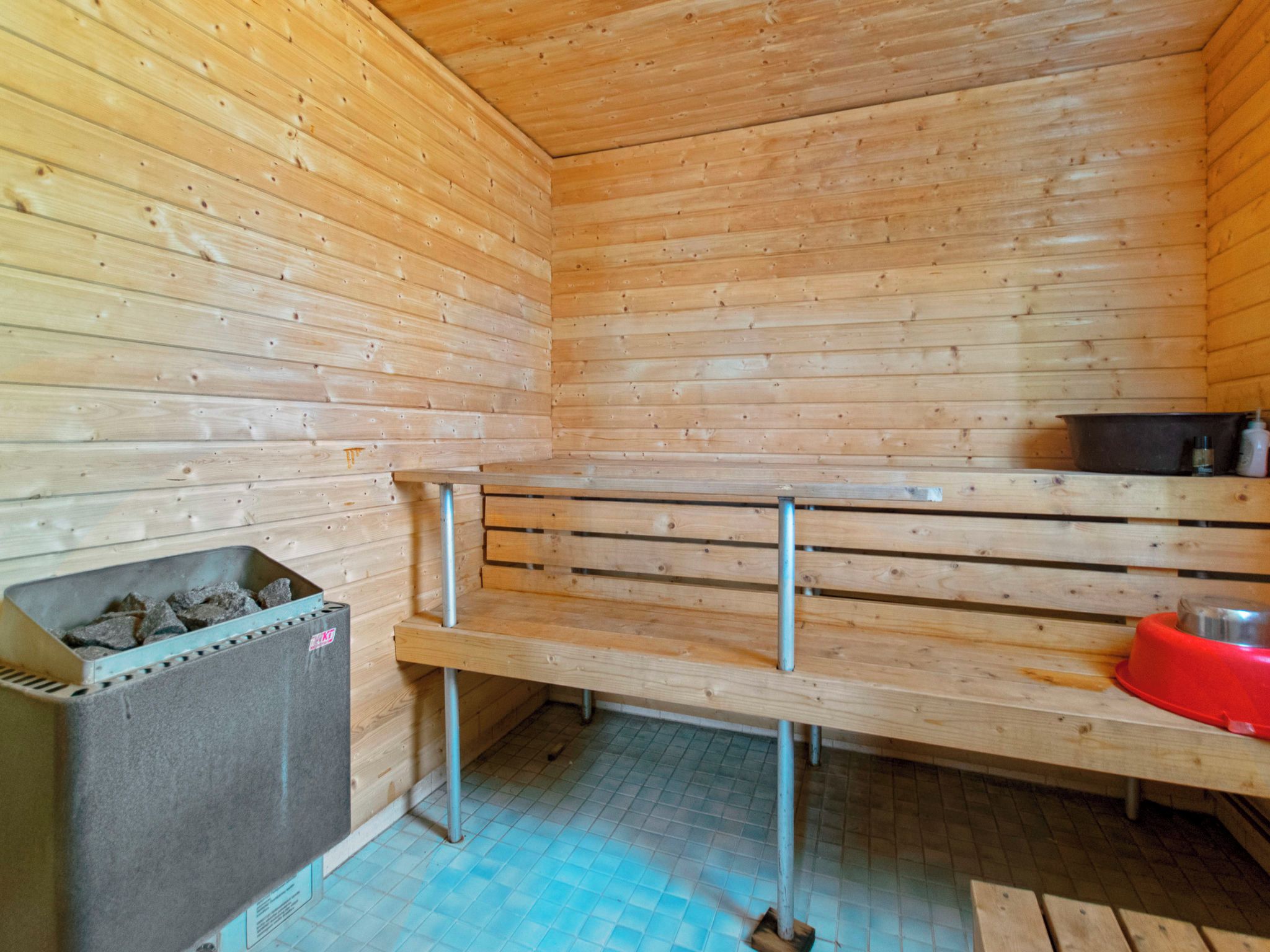Una sala de sauna con paredes de madera, bancos, un banco de metal, un cubo de basura y una cuenca roja.