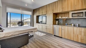 Ein modernes Schlafzimmer mit Bett, kleinem Nachttisch und Blick auf einen Berg durch große Fenster. Daneben befindet sich eine kompakte Küche mit Holzschränken, einer Mikrowelle und einem kleinen Kühlschrank.