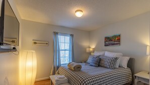 Een netjes ingerichte slaapkamer met een bed, bedlampjes, een nachtkastje met handdoeken, een staande lamp en een raam met gordijnen.