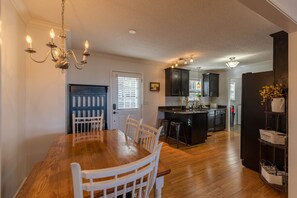A dining area with a wooden table and chairs, a chandelier, and a kitchen in the background.