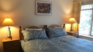 Ein Schlafzimmer mit einem Bett, zwei Nachttischen, zwei Lampen, einem gerahmten Bild an der Wand und einem Fenster mit Vorhängen.