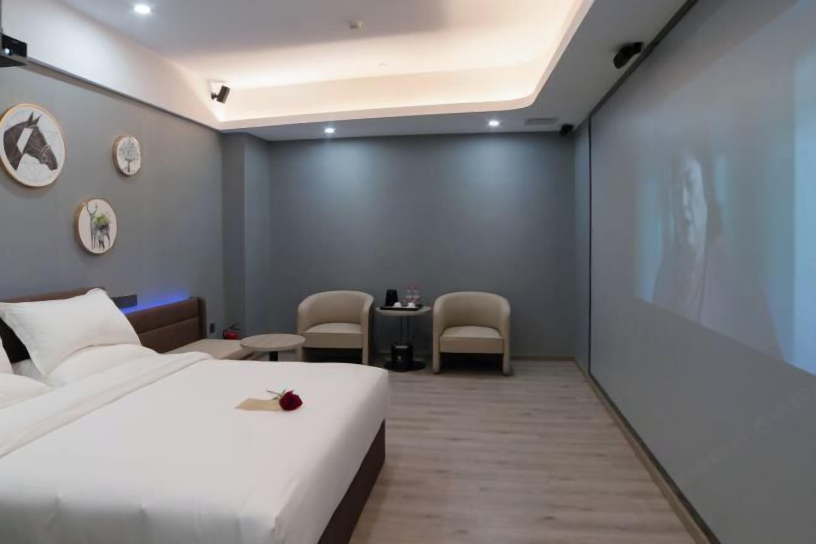 Une chambre d’hôtel moderne équipée d’un lit, de deux fauteuils, d’une petite table et d’un écran de projection fixé au mur.