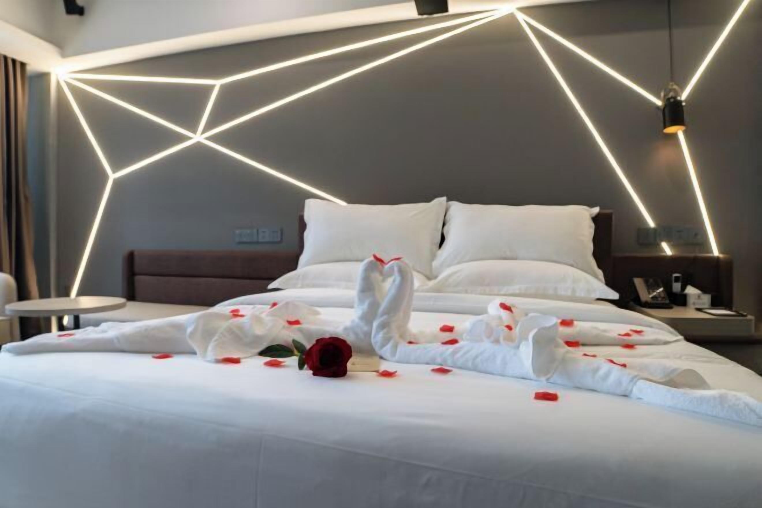 Una habitación de hotel moderna con una instalación de luces geométricas en la pared, una cama bien hecha con sábanas blancas y pétalos de rosa, y una sola rosa roja sobre la cama.