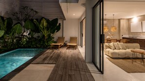 Un espace piscine intérieur moderne, doté d’un plancher en bois, de chaises longues et d’une cuisine visible à travers des portes vitrées.